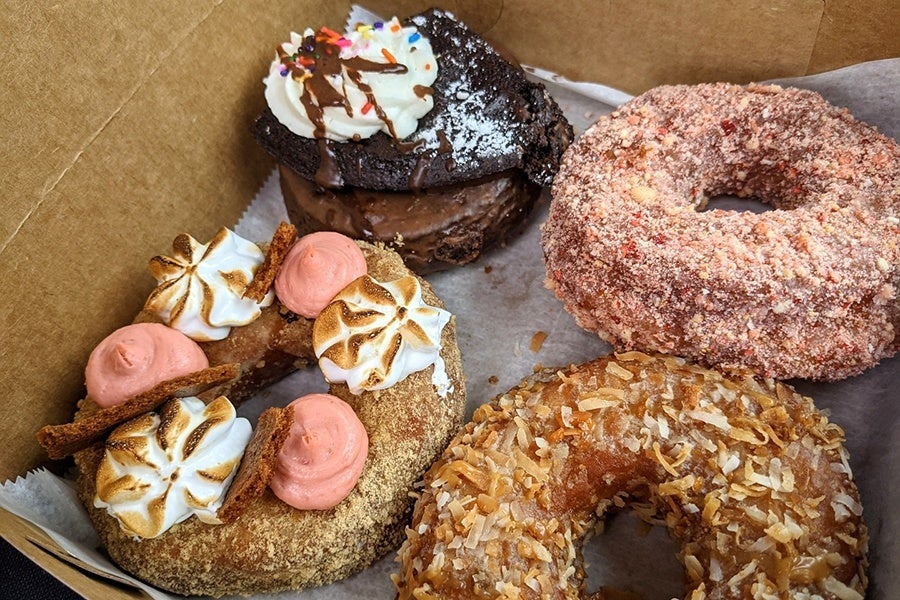 Four different doughnuts sit in a cardboard box with a variety of decadent toppings.