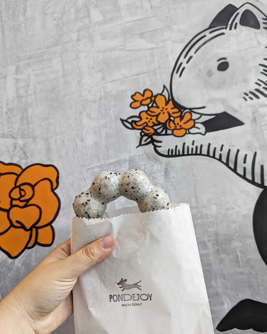 A hand holds up a paper bag with mochi doughnut coming out of it, shaped like a ring of bubbles.