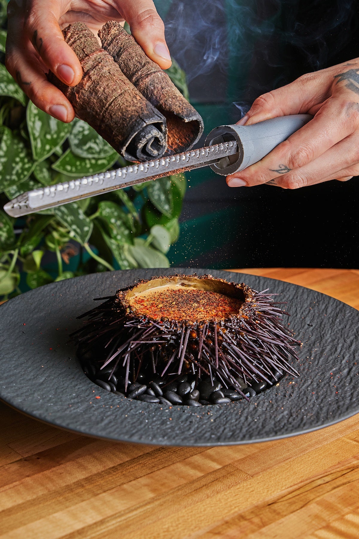A hand grates cinnamon over sea urchin, split in half on a black plate.