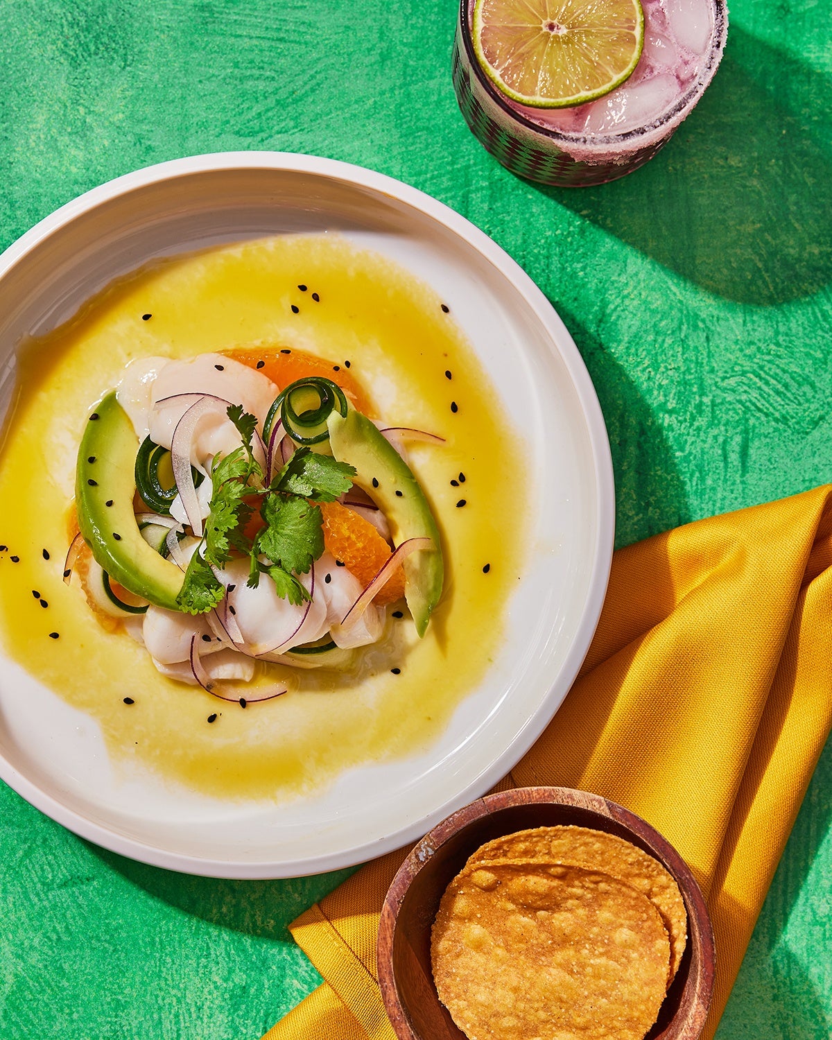 Overhead view of a bright aguachile with avocado and a yellow sauce. A pink cocktail sits nearby.