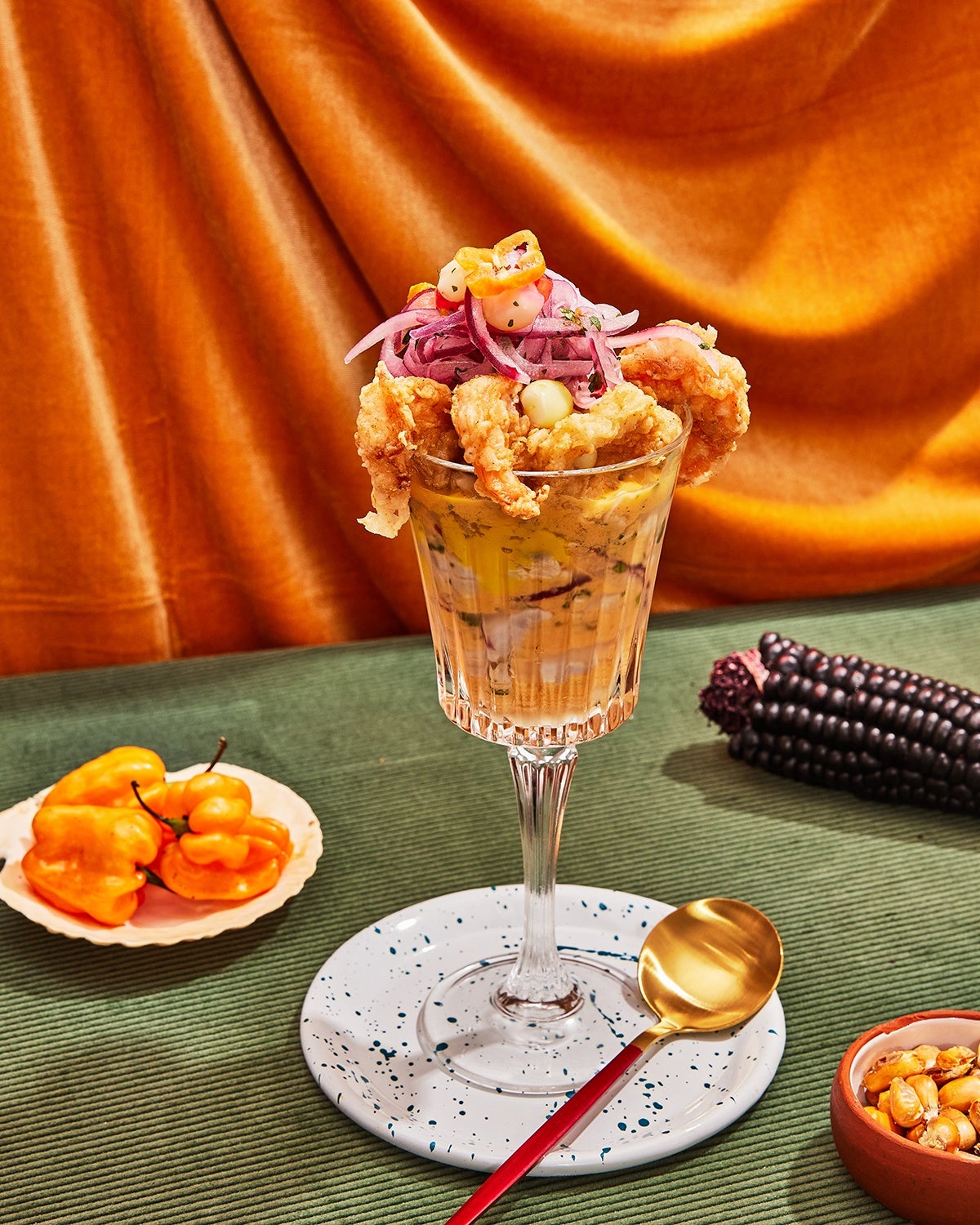 A tall glass of ceviche with shrimp sitting on top is displayed on a green surface in front of a velvety orange drape, with accoutrements surrounding it, including an ear of purple corn.