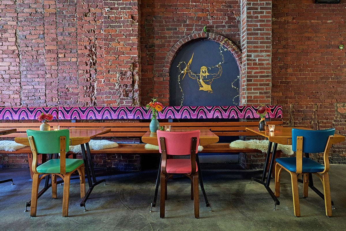 Photo of the interior of Asta restaurant, which shows a brick wall with a painting of Zeus, three two-top tables with flowers, and three colorful chairs.