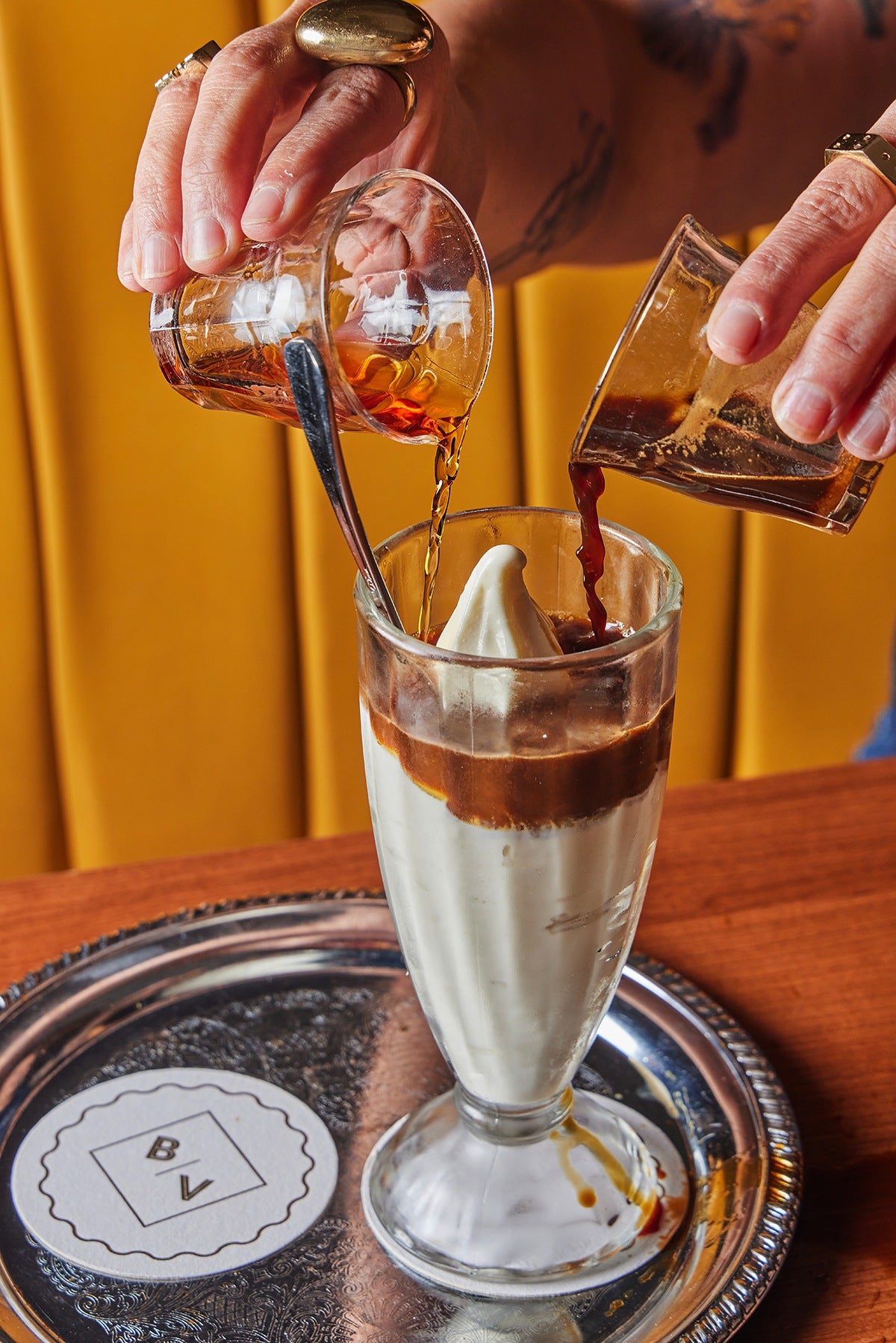 Two hands pour two different liquids—one a red booze, and one coffee—over an elegant glass of soft-serve vanilla ice cream.