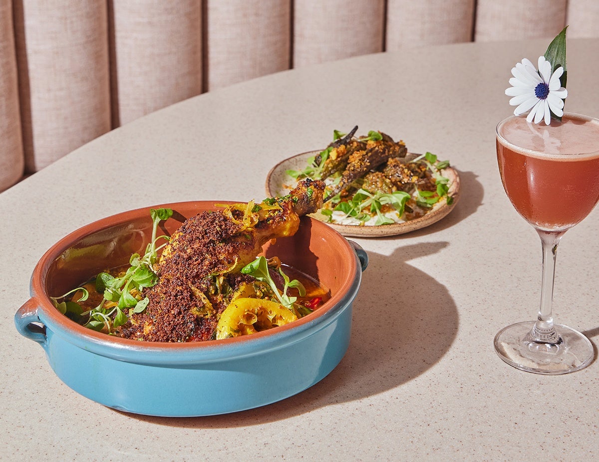 A shallow blue pot of seared okra and yassa chicken, accompanied by a brown rum-based cocktail with a white and blue daisy-like flower dipped into the drink.