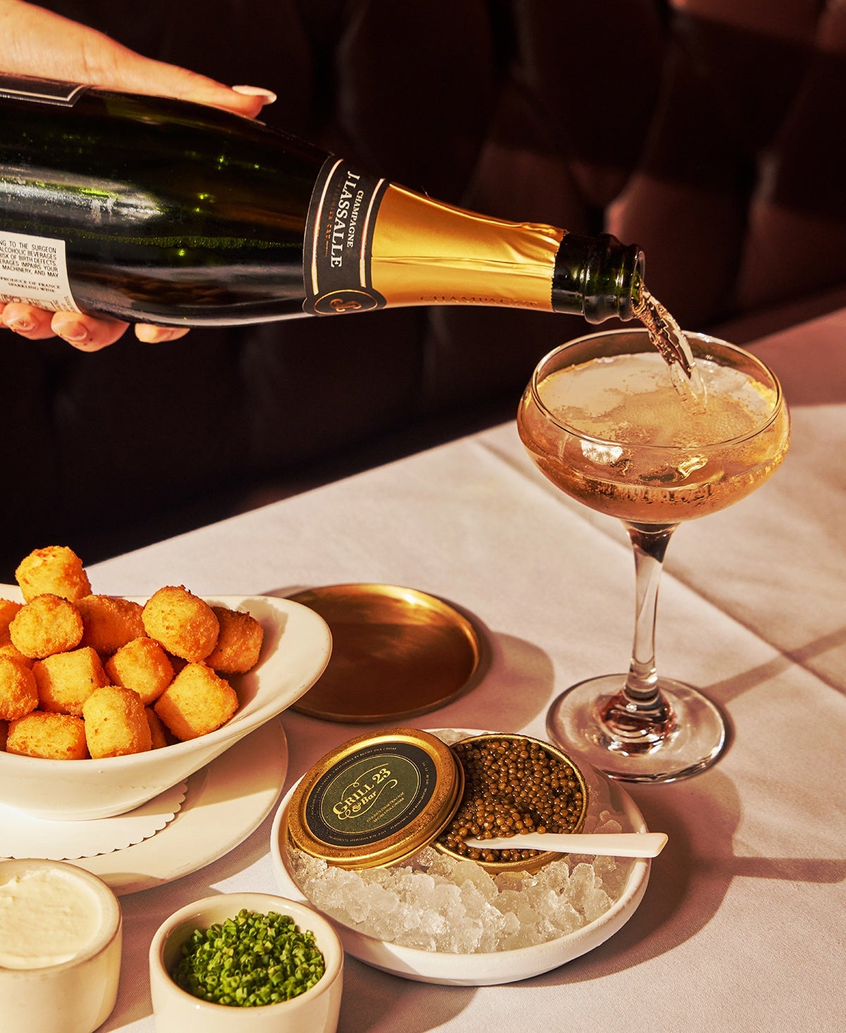 A close-up of a hand pouring champagne into a glass, over a white tablecloth setting of a caviar tin on ice and a bowl of lightly fried tater tots.