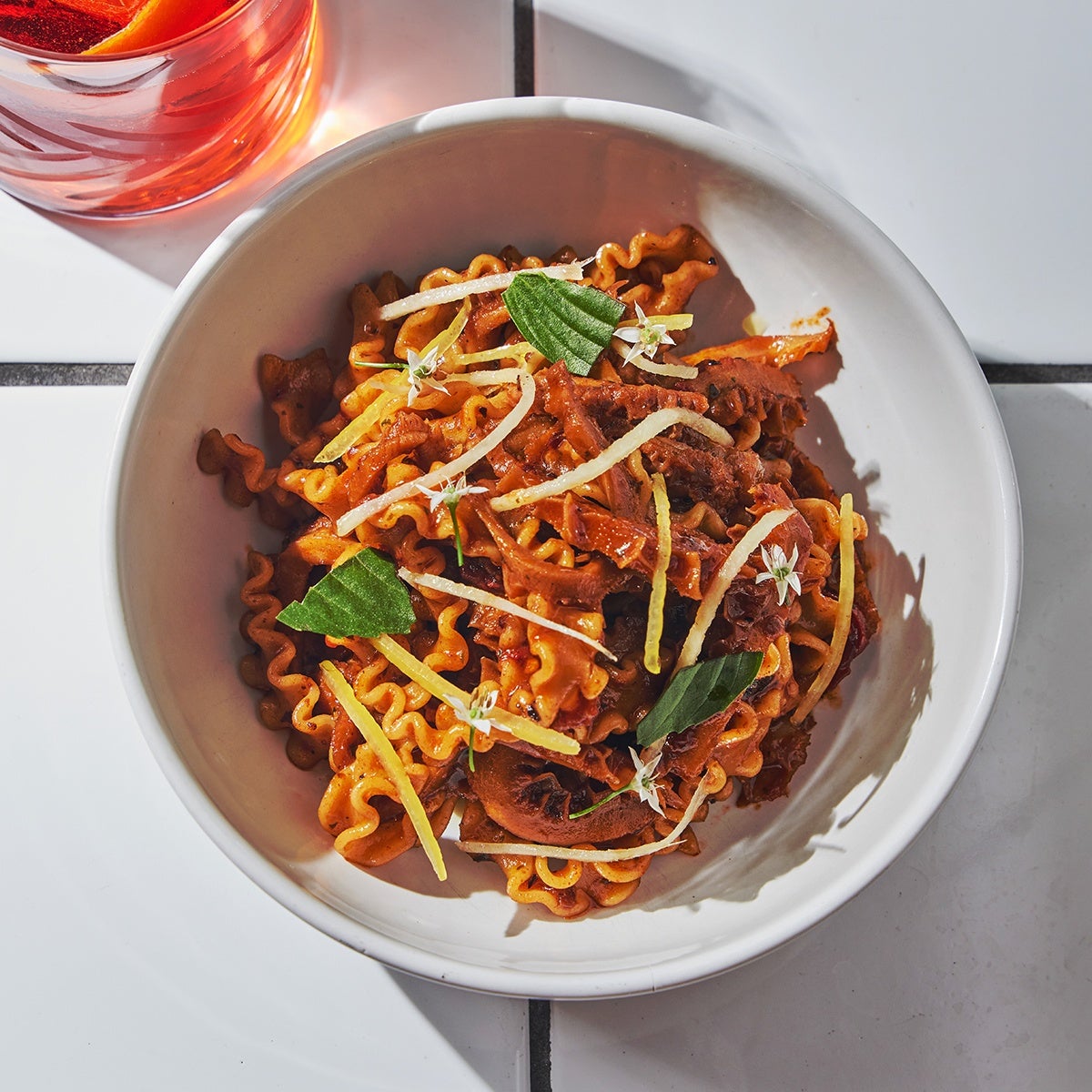 Overhead view of a pasta dish featuring thick noodles with a ruffled edge in a white bowl on a white tiled surface.