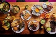 Overhead view of a wooden table covered with food, including samosas, a mango salad, grilled chicken thighs, and more.