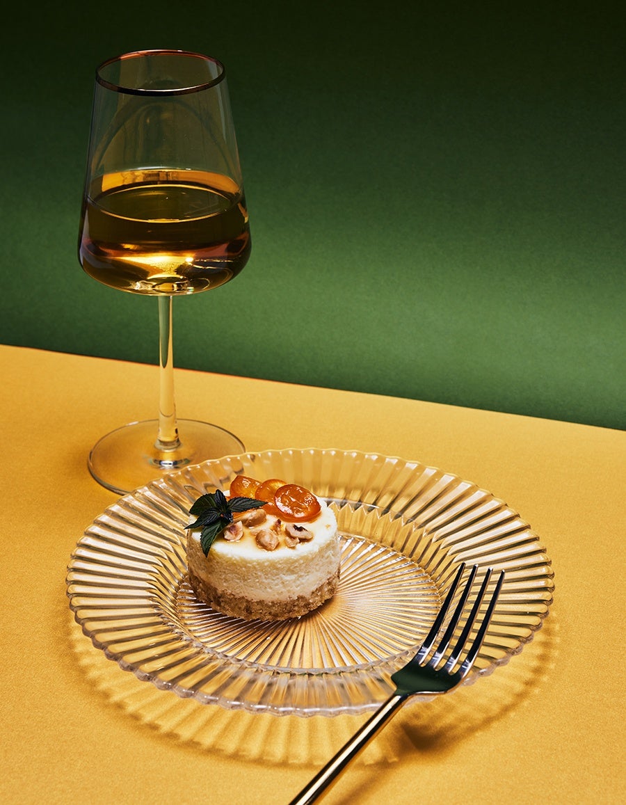 An elegant round dessert topped with candied kumquats is displayed on a glass plate with a wine glass on the side.