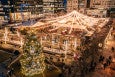 An outdoor holiday market in the middle of the city features lots of white lights and lots of people dressed in warm clothes. There's a big Christmas tree and curling lanes visible.