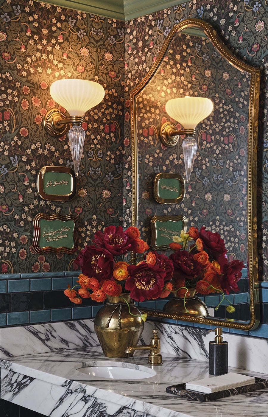 An elegant restaurant bathroom features delicate floral wallpaper and a brass vase full of fresh red and orange flowers.