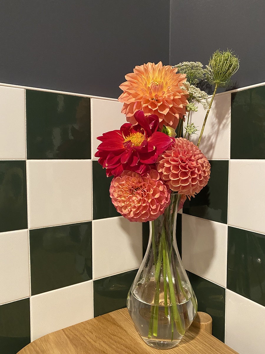 Closeup of a black-and-white tiled wall in a restaurant bathroom, with a glass vase of pink, red, and orange flowers sitting on a wooden surface.