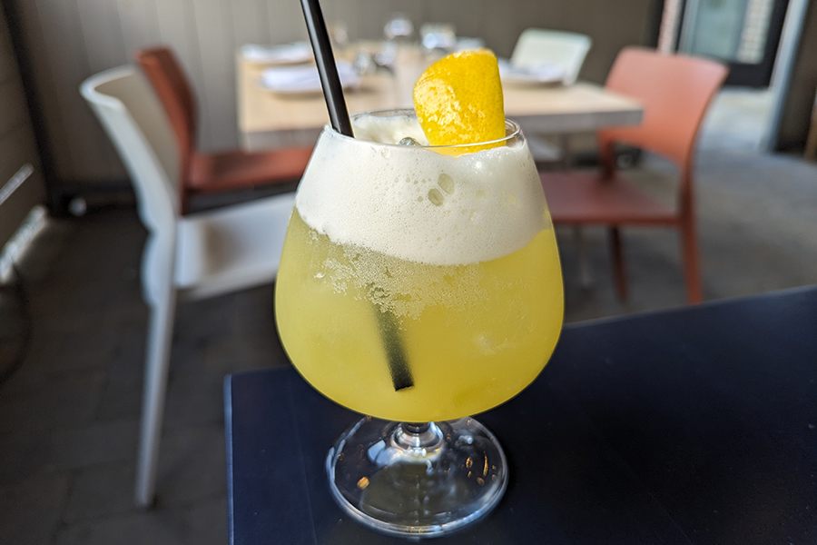 A fizzy yellow drink with a lemon wedge sits on a table on a restaurant patio.