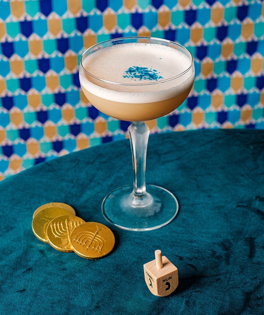 A pale pink cocktail with foam and blue sprinkle garnish sits on a blue surface, surrounded by Hanukkah gelt and a dreidel.