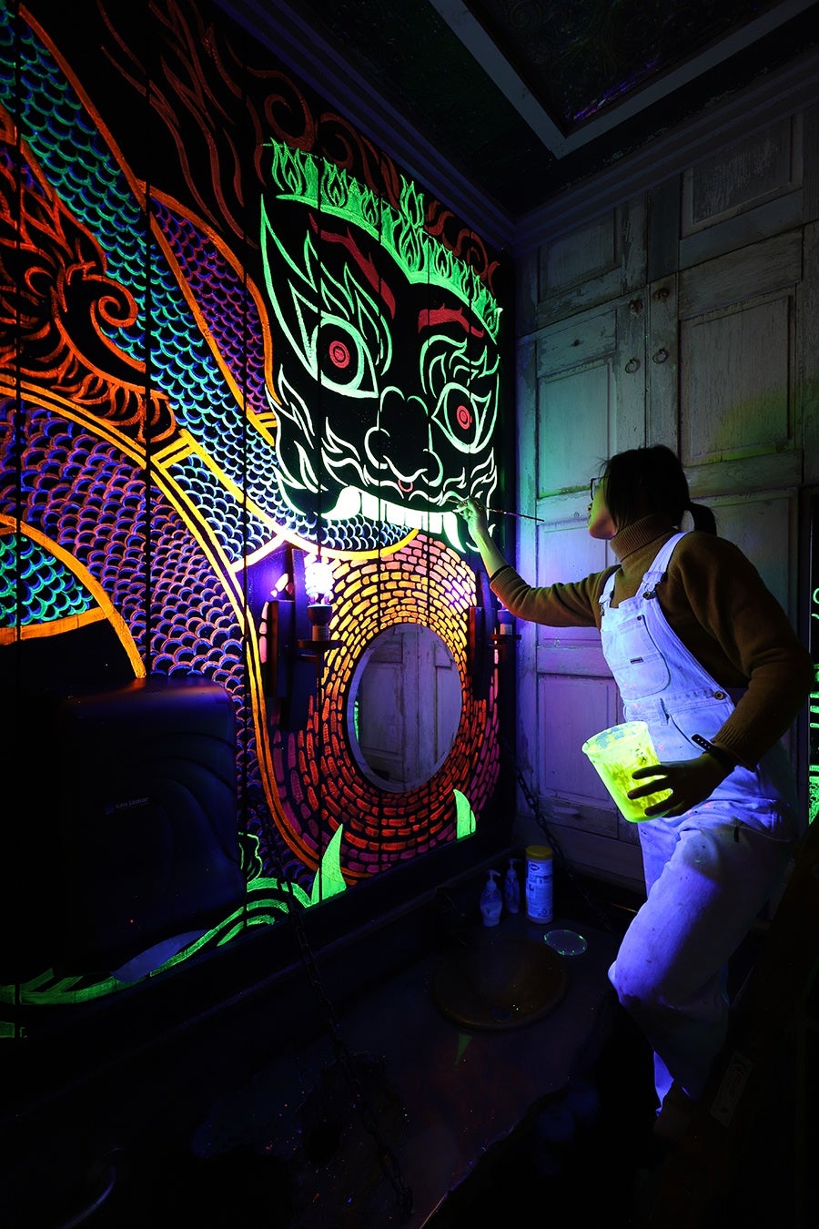 A person in overalls paints a blacklight mural of a Thai dragon in a small, dark room.