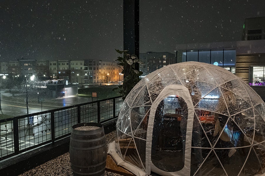 A see-through dining igloo sits atop a restaurant rooftop with city skyline views as snow falls.