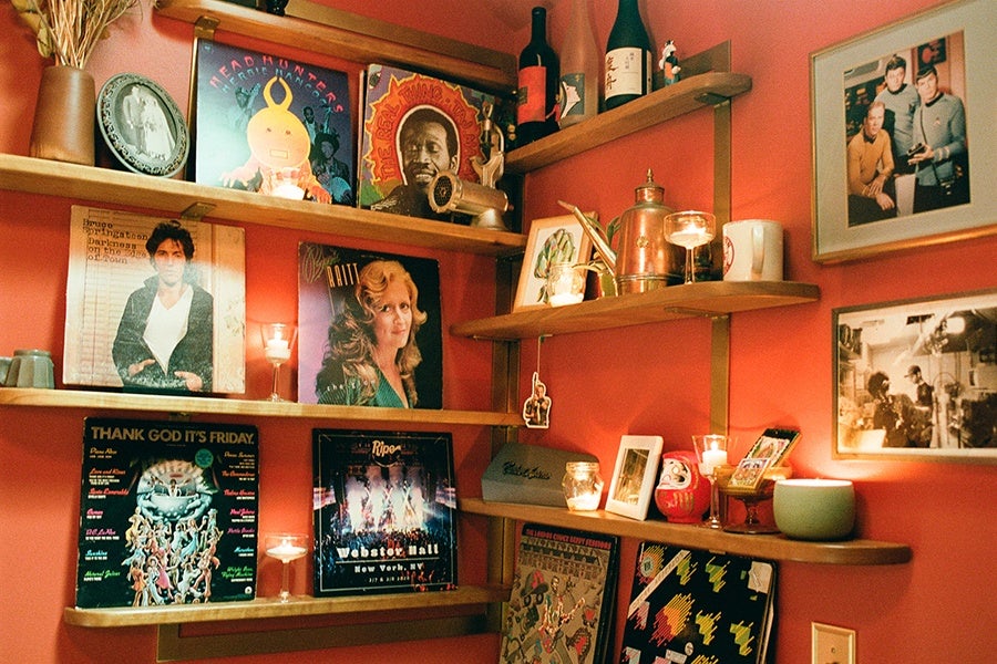 A restaurant bathroom has orange walls and wooden shelves covered with album covers and a variety of knick-knacks.