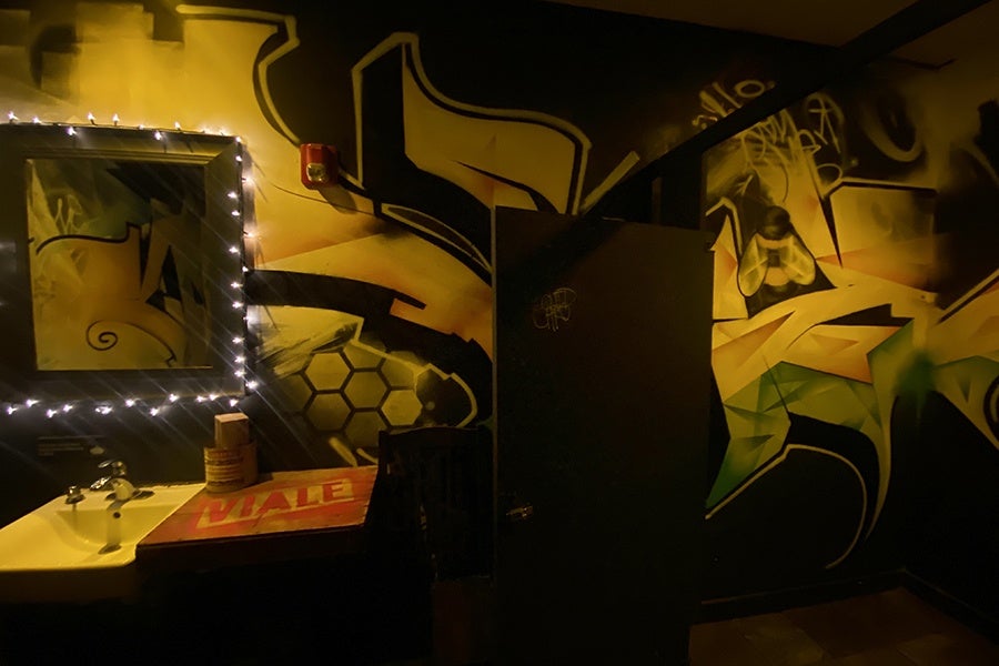 A dark restaurant bathroom is covered with graffiti-style art. White string lights surround the mirror over the sink.