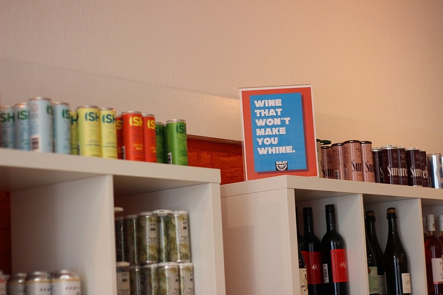 Bottled and canned nonalcoholic wine lines several shelves in a store, alongside a sign that says wine that won't make you whine.