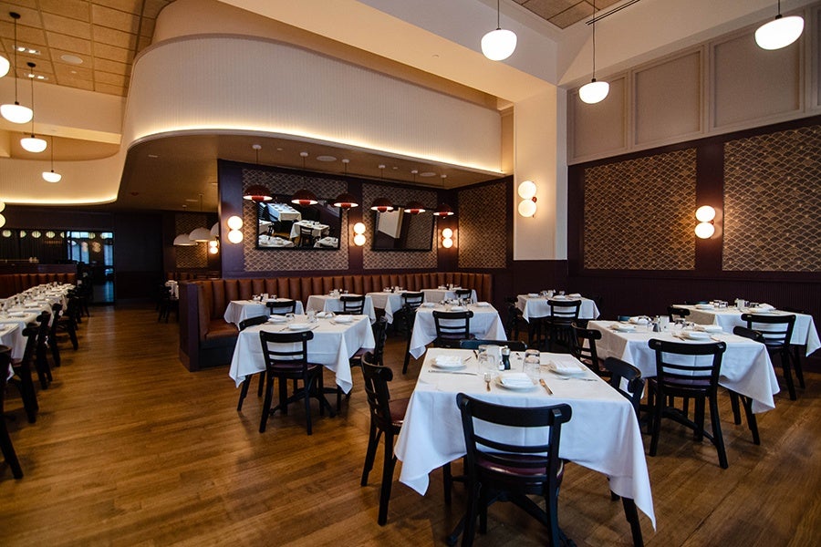 A restaurant dining room features white tablecloth-covered tables and brown banquettes.