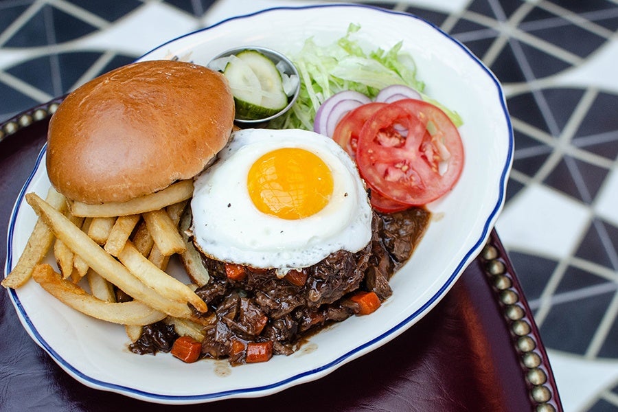 A burger is topped with a runny egg and a meaty gravy, with fries, tomatoes, onions, and lettuce on the side.