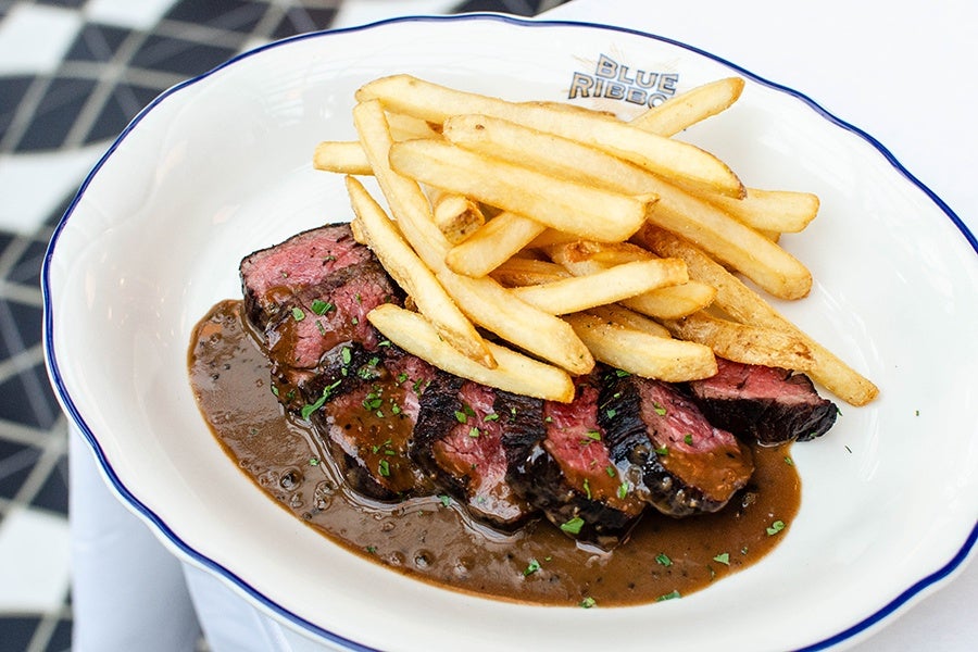 Rare sliced steak is served with fries on a plate that says Blue Ribbon atop a white tablecloth.