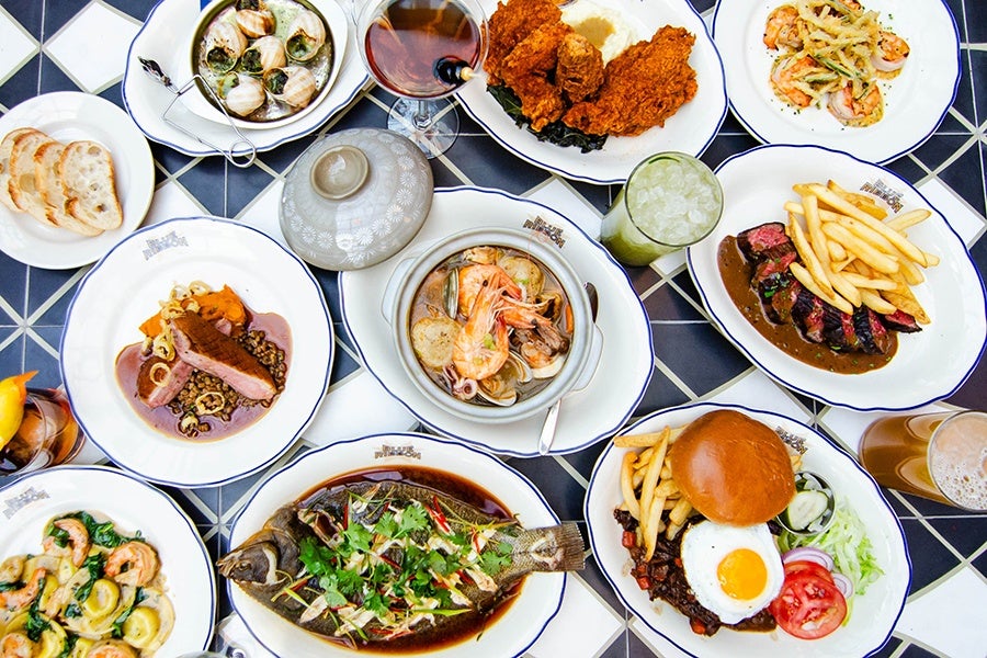 An overhead view of around 10 dishes of food, including steak frites, a burger, a whole fish, fried chicken, and more.