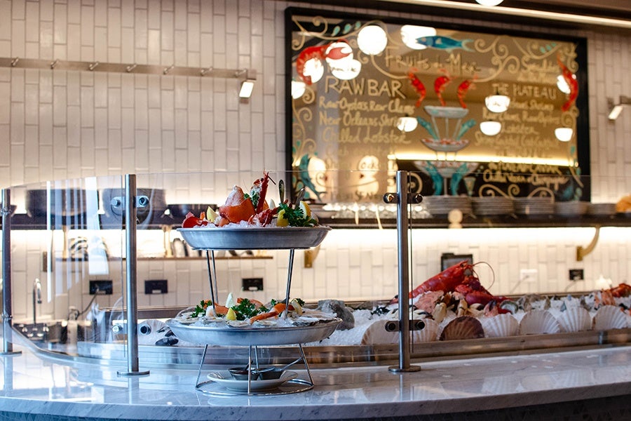A two-tier seafood tower sits on a white marble raw bar with a menu on the wall painted on a large mirror.