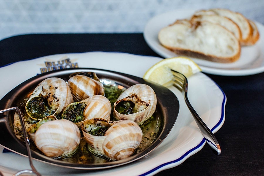 A silver plate of parsely-and-butter-soaked snails is accompanied by a lemon wedge and a few slices of a baguette.