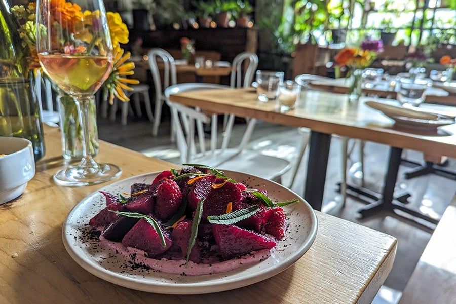 A plate of raspberries, beets, and herbs sits on a table in a restaurant full of flowers and light wooden tables.
