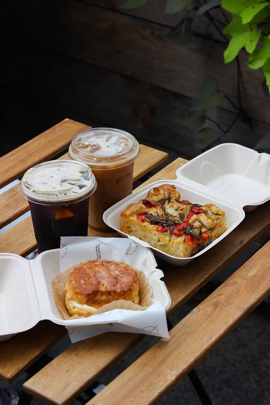 Two iced coffees sit on a small outdoor dining table with takeout containers of a biscuit sandwich and a thick slice of focaccia.