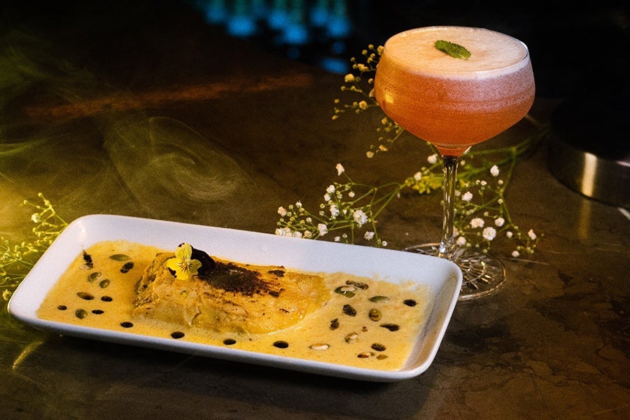 A tamale is smothered in a yellow cream sauce and accompanied by a pink cocktail.