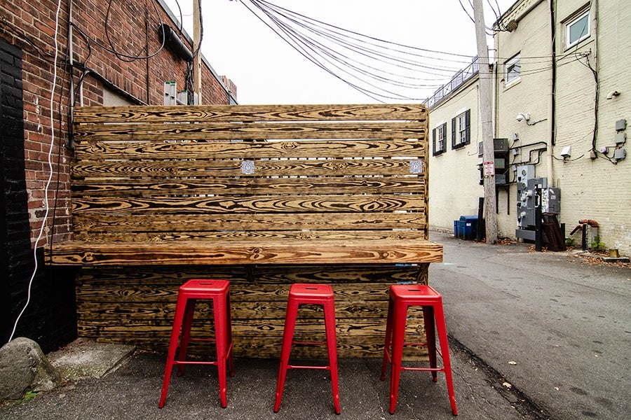 Three red stools sit in front of a wooden counter against a brick building in a wide alley.