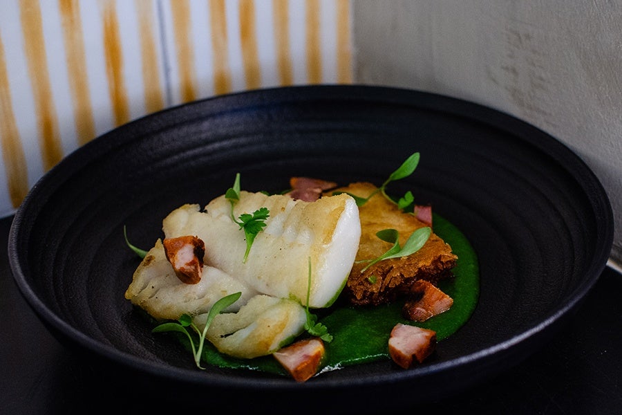 A seared filet of cod leans on a crispy rectangle of potato, with chunks of sausage in a pool of green sauce.