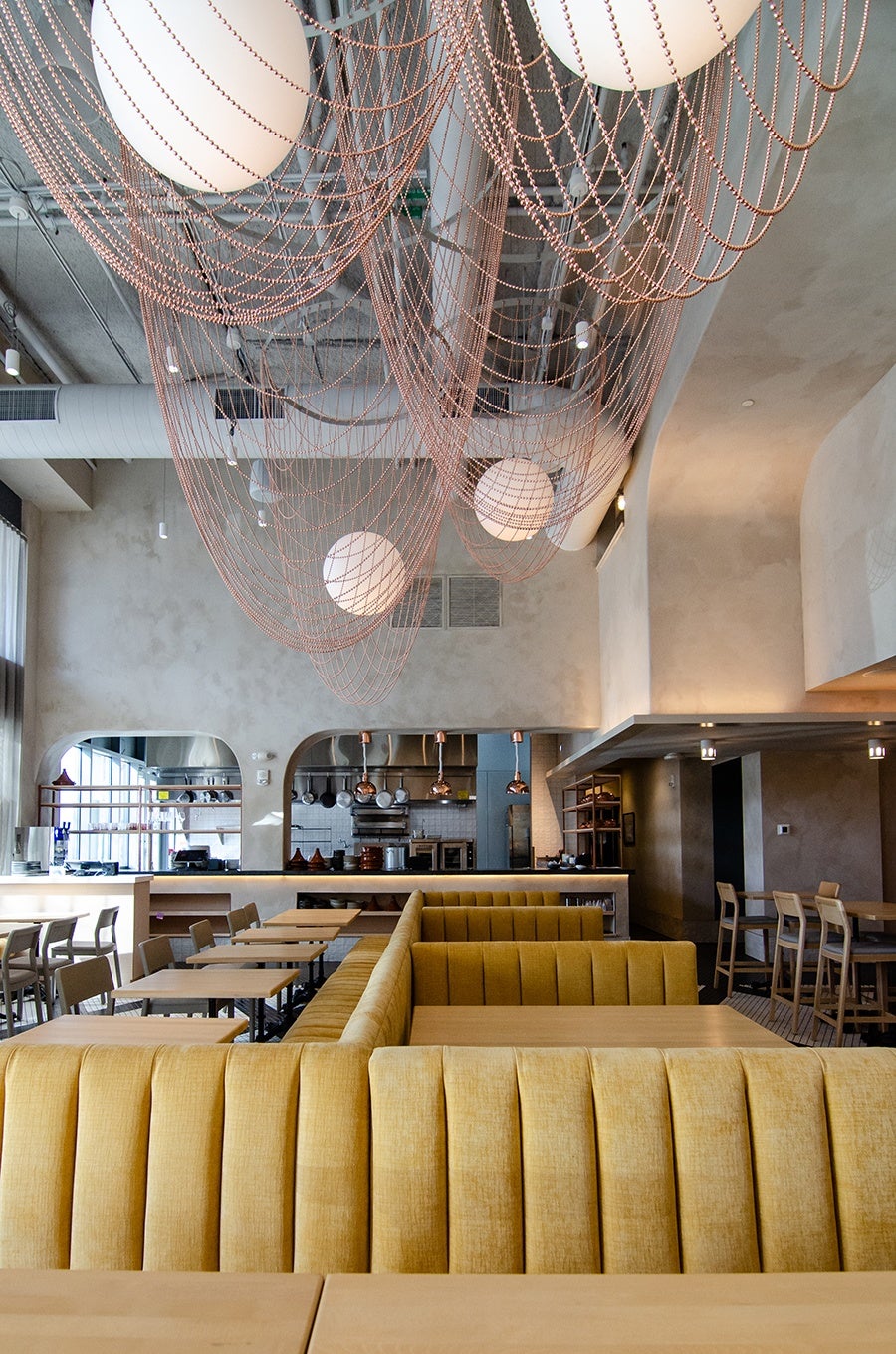 An empty restaurant space features pale yellow banquettes, light limewashed walls, and large spherical lights surrounded by big loops of copper beads.