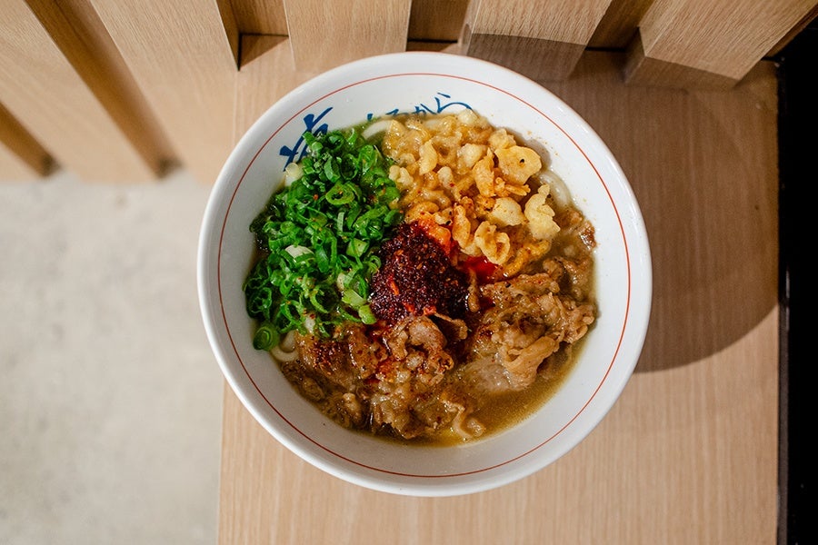Overhead view of a bowl of udon with thinly sliced beef, scallions, crispy tempura flakes, and chili oil.