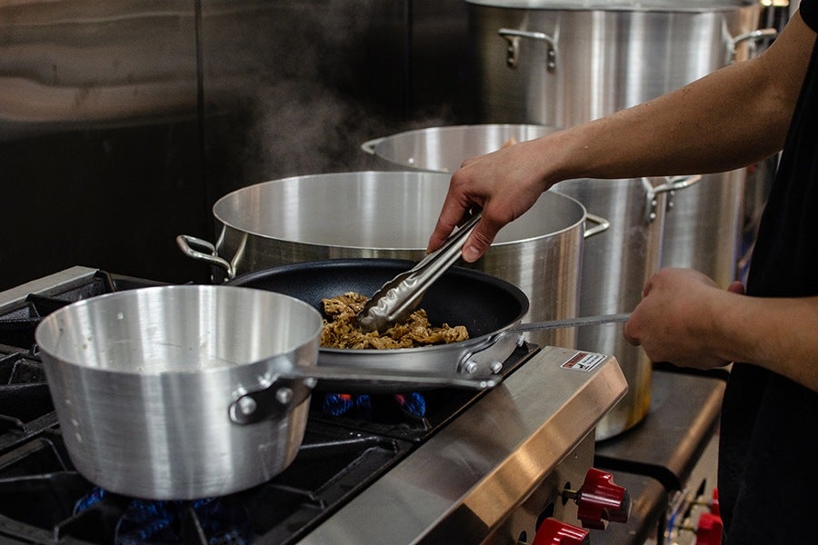 A chef holding tongs cooks thinly sliced beef in a pan on a commercial stovetop.