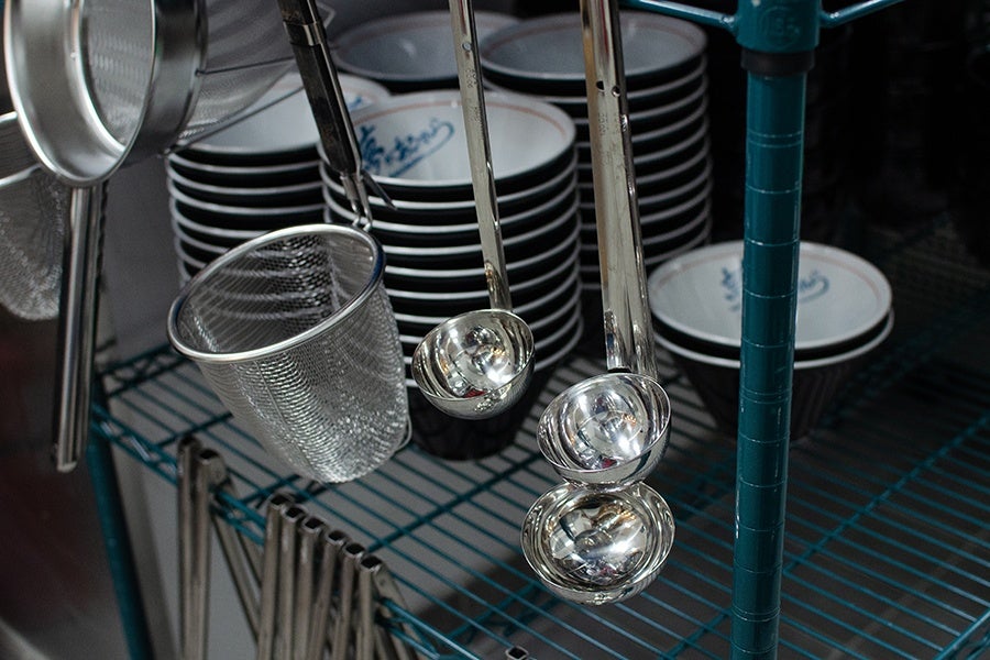 Ladles, strainers, and tongs hang from the edge of restaurant kitchen shelving, which holds stacks of large soup bowls.