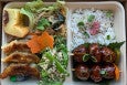 Overhead view of a bento box with seasoned white rice, saucy meatball skewers, gyoza, noodles, butter cake, and more.