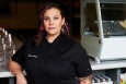 A woman in a black chef's shirt and pink streaks in her hair poses for a headshot.