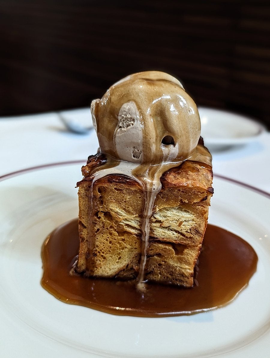 A tall slice of bread pudding sits in a pool of caramel and is topped with a melty scoop of ice cream.