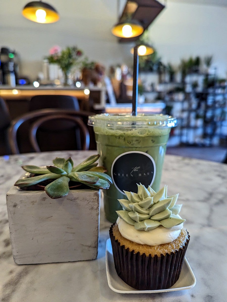 A cupcake with frosting in the shape of a succulent sits next to an actual succulent and a matcha latte on a white marble table.