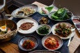 A table is covered with dishes of Korean food, including a selection of small side dishes of kimchi and more.