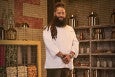 A man with a full beard and long dreads wears a white chef's coat with Top Chef branding and stands smiling for a portrait in a large pantry.