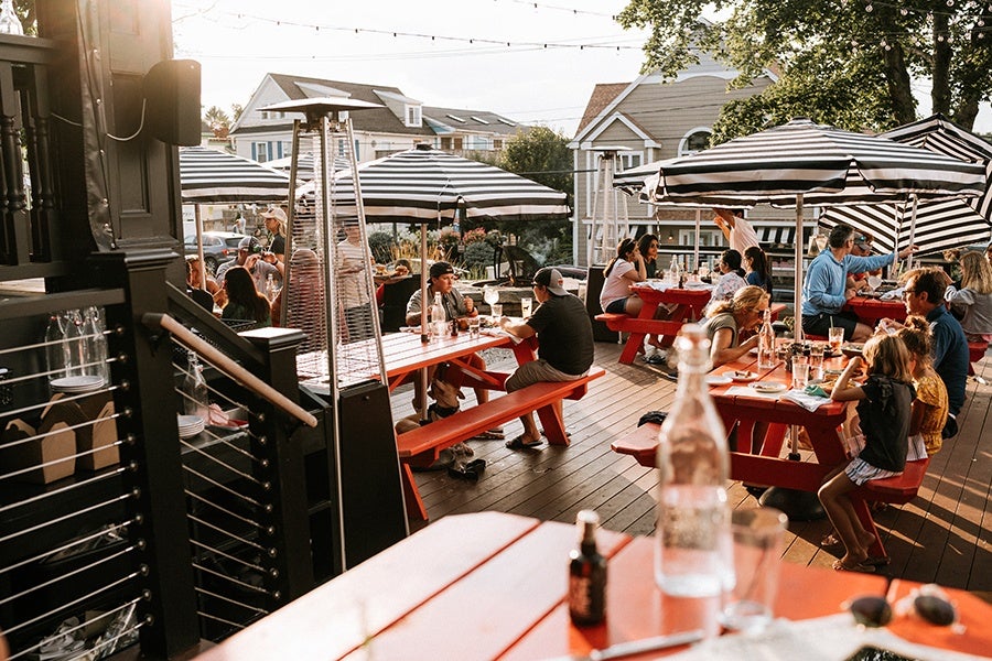 People in casual summer clothing sit at orange picnic tables on the sunny patio of a brewery.