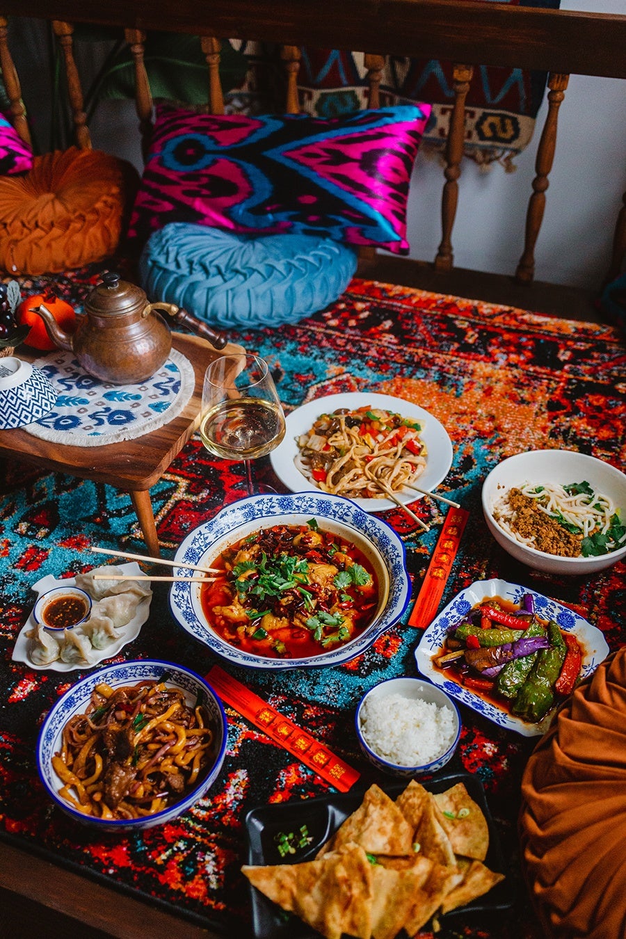 Several Uyghur noodle dishes and a dish with hot chili oil are spread on a colorful carpet with a copper pot of tea and a glass of wine in view.