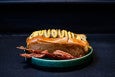 A hot dog is topped with a squiggle of a mustard-like sauce and has a side of crispy bacon strips.