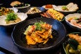 Thai-style small plates are spread across a table, including sausage in a bun, dumplings, noodles in a thick orange sauce, and more.