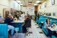 Patrons eat inside an old-fashioned diner with teal booths and stools at a counter in front of an open kitchen.