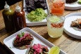 Tacos, colorful cocktails, and guac with blue corn chips sit on a wooden table.