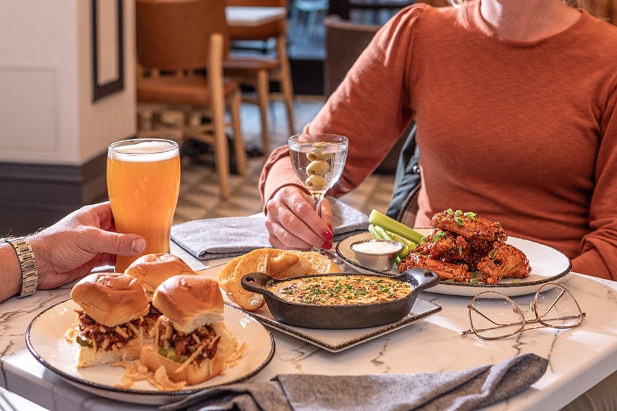 Two people sit at a restaurant table eating sliders and wings, one drinking beer and one drinking a cocktail.